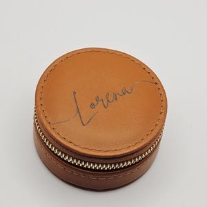 May include: A round, brown leather jewelry case with a gold zipper. The word "Lorena" is written in cursive on the top. The case is closed and sits on a white surface.