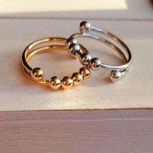 May include: Two open-band rings, one gold-toned and one silver-toned, each featuring a series of small, movable metal beads along the front section. The rings are circular with a gap, designed with a smooth, polished metallic finish.