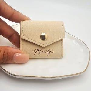 May include: A small, beige suede pouch with a flap closure and a gold-colored snap. The name "Marilyn" is elegantly printed in cursive on the front. The pouch is designed to hold small items, such as jewelry.