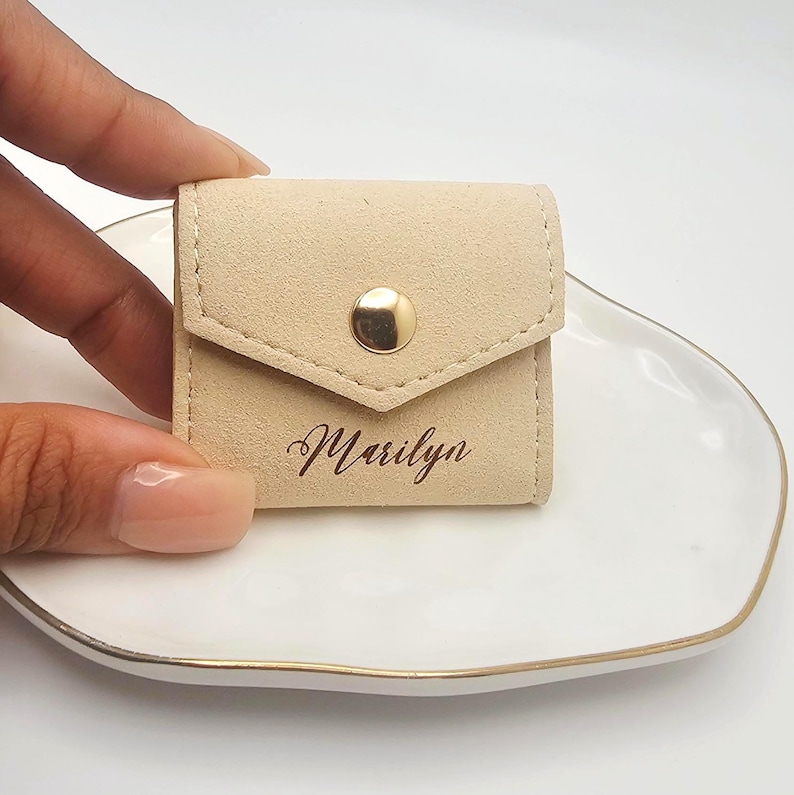 May include: A small, beige suede pouch with a flap closure and a gold-colored snap. The name "Marilyn" is elegantly printed in cursive on the front. The pouch is designed to hold small items, such as jewelry.