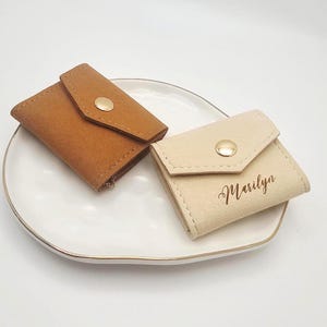 May include: Two small, suede-like pouches with flap closures and gold-colored snap buttons. One pouch is tan, the other is beige with the name "Marilyn" in script. The pouches are on a white plate with a gold rim.