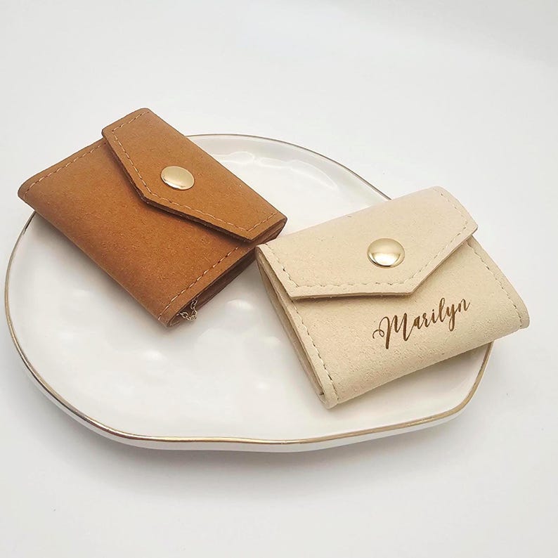 May include: Two small, suede-like pouches with flap closures and gold-colored snap buttons. One pouch is tan, the other is beige with the name "Marilyn" in script. The pouches are on a white plate with a gold rim.