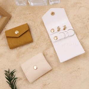 May include: An assortment of small jewelry cases in various colors. A white, snap-closure case holds earrings and rings. A small, mustard-colored case and a cream-colored case are also shown. All cases have gold-colored snap closures.
