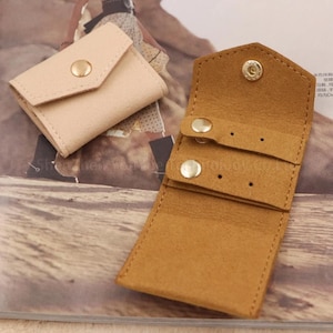 May include: Two small, open leather pouches. One is beige and closed with a gold snap. The other is open, tan, and has two straps with gold snaps, designed to hold jewelry. The pouches are on a patterned surface.