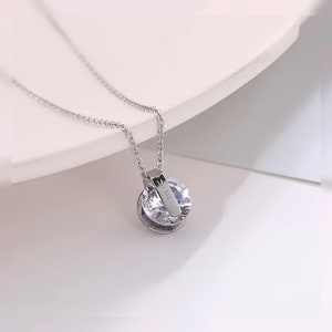 Puede incluir: Un collar de cadena de plata con un colgante redondo que presenta un cristal transparente. El colgante tiene una pequeña barra de plata con la palabra "14K" grabada en él.