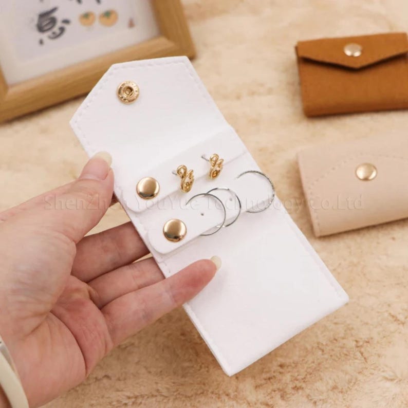 May include: A white, snap-closure jewelry organizer with gold-colored accents. The organizer holds two pairs of gold earrings and two silver hoop earrings. Additional small, tan and brown pouches are in the background.