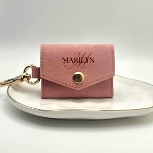 Puede incluir: Pequeño estuche de color rosa empolvado con el nombre "Marilyn" impreso. Cuenta con un cierre de botón a presión dorado y un mosquetón dorado. El estuche descansa sobre un plato blanco con borde dorado.
