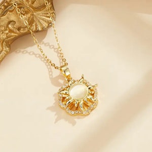 May include: Gold-toned necklace featuring a sun pendant. The pendant has a central, oval-shaped, opalescent stone surrounded by a halo of small, clear crystals and sun ray-like projections. The chain is delicate and the necklace is displayed against a neutral background.
