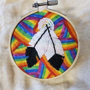 Könnte beinhalten: Stickrahmen-Kunst mit Stickerei, die einen Regenbogenhintergrund mit einem weißen und schwarzen Federdesign in der Mitte zeigt.