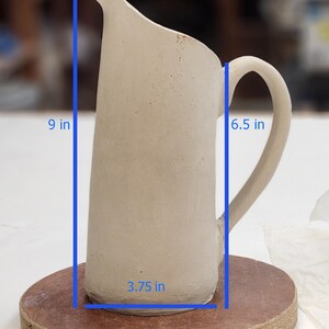 May include: A handmade, off-white ceramic pitcher with a curved handle. The pitcher is 9 inches tall, 6.5 inches wide, and 3.75 inches in diameter. The pitcher is sitting on a round, brown surface. The text "Measurements" is visible.