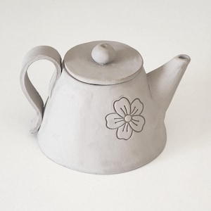 May include: A small, grey ceramic teapot with a flower design. The teapot has a lid with a knob on top and a spout on the side.