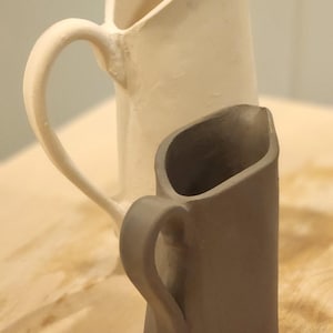 Könnte beinhalten: Zwei Keramikkrüge. Einer ist cremefarben, der andere ist ein gedämpftes Grau. Beide haben einen gebogenen Griff und einen Ausguss. Die Krüge stehen auf einer hellen Holzoberfläche.