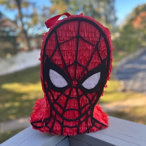 May include: A red Spider-Man piñata with black web detailing and white eye shapes. The piñata is made of red fringed paper and has a red ribbon for hanging. The background is blurred, showing a green lawn and trees.