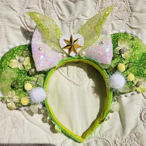 May include: Green and white Minnie Mouse ears with a sparkly bow, a gold star, and white pom-poms. The ears are decorated with green leaves and white flowers.