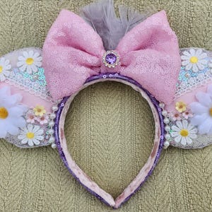 May include: Pink and white mouse ears with a pink bow, purple headband, and white daisy embellishments.