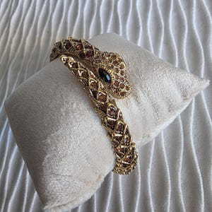 Op de afbeelding: Een goudkleurige armband in de vorm van een slang met een gestructureerd, schubachtig ontwerp ingelegd met kleine, glinsterende bruine kristallen. De slangenkop heeft een opvallend, glad zwart stenen oog en het lichaam is spiraalvormig.