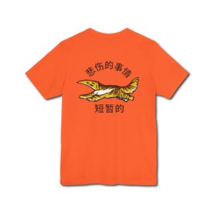 Peut inclure: T-shirt orange vif avec un motif d'une créature aviaire jaune et marron, accompagné de caractères chinois. Le col est rond et les manches courtes. Vêtement décontracté.