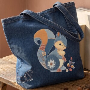 May include: A dark blue denim tote bag featuring a large, multi-textured patch of a squirrel with a bushy tail. The squirrel design incorporates floral patterns and soft earth tones. A smaller, subtle line-art squirrel graphic is printed near the base.