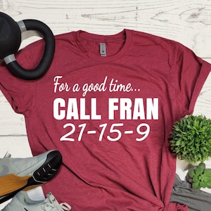 May include: A red t-shirt with the text "For a good time... CALL FRAN 21-15-9" printed on it. The shirt is paired with gray athletic shorts and a pair of gray and black running shoes.