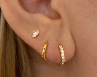 May include: Close-up of an ear adorned with three gold earrings. A small stud earring with a floral design is at the top. Below, a plain gold hoop and a gold hoop with small, clear stones are visible. The earrings are set against a background of blonde hair.