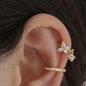 May include: A close-up of an ear adorned with multiple pieces of gold-colored jewelry. The jewelry includes a floral ear cuff, a hoop, a bar with a single stone, a crescent moon stud, and a small round stud. The text "Amygrace Studio" is visible.