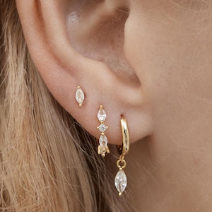 Könnte beinhalten: Eine Sammlung goldener Ohrringe, darunter kleine Ohrstecker und Creolen mit tropfenförmigen Edelsteinen. Die Ohrringe sind vor dem Hintergrund eines Ohrs und blonden Haaren abgebildet.