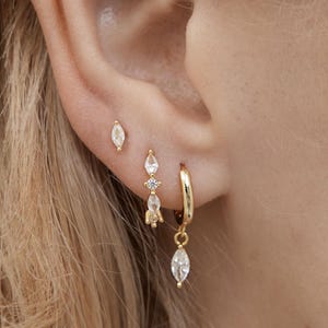 Puede incluir: Primer plano de una oreja adornada con tres juegos de pendientes dorados. El primer juego es un pequeño pendiente de botón ovalado. El segundo juego presenta un diseño colgante con un pequeño diamante. El tercer juego es un aro pequeño con una gema ovalada colgante.