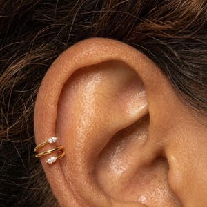 May include: Close-up of an ear adorned with a gold ear cuff. The ear cuff features a double-band design with two small, sparkling gemstones. The jewelry is set against the backdrop of the ear's natural skin tone. The text "Amygrace Studio" is visible.