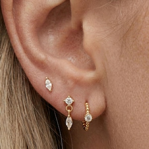 May include: Close-up of an ear adorned with three sets of gold earrings. One is a marquise-shaped stud, another features a diamond stud with a dangling marquise gem, and the third is a small hoop with a single diamond.