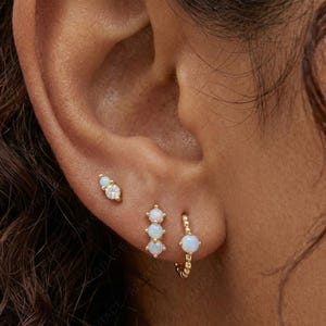 May include: A close-up of an ear adorned with three sets of earrings. The earrings are gold-toned and feature opal and diamond accents. The text "Amygrace Studio" is visible at the bottom of the image.