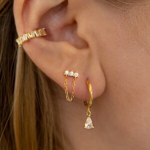 Könnte beinhalten: Goldfarbenes Ohrschmuck-Set. Enthält einen Ohrklemme, einen Ohrstecker mit Kette und einen Creolen-Ohrring mit tropfenförmigem Anhänger. Der Schmuck ist mit klaren, funkelnden Steinen besetzt. Das Ohr steht im Fokus vor unscharfem Hintergrund.