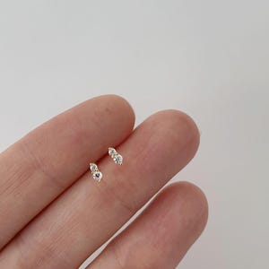 May include: A pair of small gold-colored stud earrings, each featuring two clear, round gemstones. The earrings are displayed on a light background. The jewelry is delicate and minimalist.