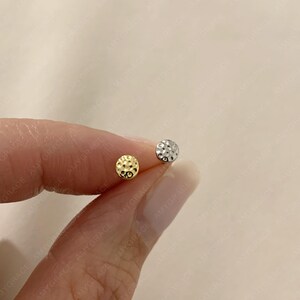 Puede incluir: Un par de pequeños pendientes de botón. Uno es de color dorado con una superficie texturizada, y el otro es de color plateado con un diseño de textura similar. Los pendientes se sostienen entre los dedos. El texto "Amygrace Studio" está en la parte inferior.