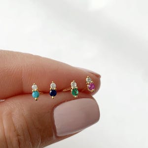 May include: Four pairs of gold stud earrings, each featuring a small gemstone paired with a clear stone. The gemstones are turquoise, navy blue, emerald green, and pink. The earrings are displayed against a white background.
