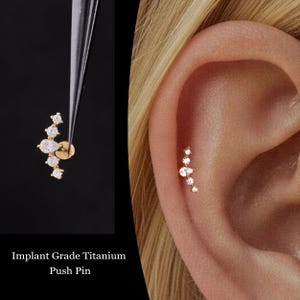 May include: A gold-colored, curved ear piercing with multiple clear, sparkling gemstones. The text "Implant Grade Titanium Push Pin" is visible at the bottom of the image. The earring is shown next to an ear, demonstrating its placement.