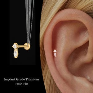 May include: Gold-colored push pin earring with two clear, sparkling gemstones. The earring is shown next to an ear piercing. The text "Implant Grade Titanium Push Pin" is at the bottom of the image.