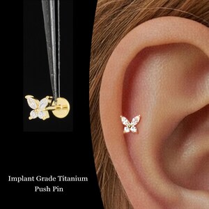 May include: A gold-colored butterfly push pin earring with clear, sparkling stones. The earring is shown in the ear and being held by tweezers. The text "Implant Grade Titanium Push Pin" is at the bottom of the image.