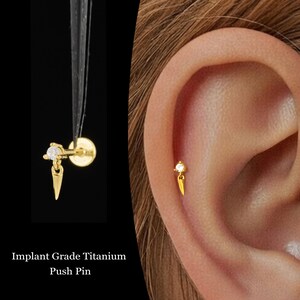 Puede incluir: Un pendiente de botón de color dorado con una pequeña gema transparente y un dije colgante puntiagudo. El pendiente se muestra en dos vistas: sostenido por pinzas y usado en una oreja. El texto en la imagen dice "Implant Grade Titanium Push Pin."