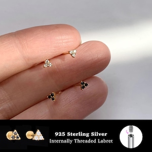 May include: Four gold-tone labret earrings featuring triangular clusters of three small stones. Two earrings have clear stones and two have black stones. The text 925 Sterling Silver Internally Threaded Labret is visible.