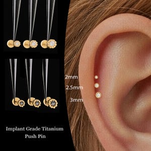 May include: Gold-colored push pin earrings with clear and black gemstones, displayed with tweezers. The image shows the earrings in various sizes: 2mm, 2.5mm, and 3mm, next to an ear with the earrings in place. Text reads "Implant Grade Titanium Push Pin."