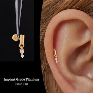May include: A gold-colored, implant-grade titanium push pin earring with two clear, sparkling gemstones. The earring is shown in two views: held by tweezers and worn on an ear. The text "Implant Grade Titanium Push Pin" is at the bottom.