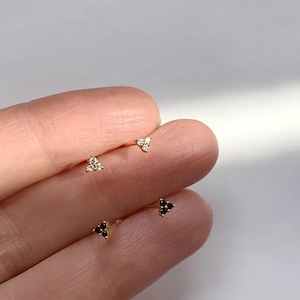 May include: A close-up of four small gold stud earrings. Two pairs of earrings are visible. One pair features three clear, sparkling stones in a triangular arrangement. The other pair has three black stones in a triangular shape.