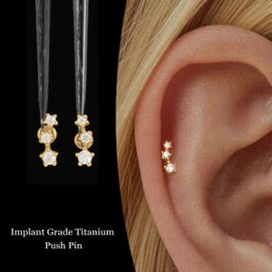 Puede incluir: Pendientes dorados en forma de estrella, cada uno con tres piedras claras y brillantes. Uno de los pendientes está en una oreja, el otro cuelga. El texto dice "Implant Grade Titanium Push Pin".