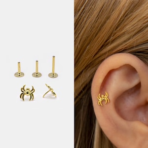 May include: A gold-toned spider-shaped stud earring with a flat back post. The set includes three different lengths of flat back posts and one spider charm top. The spider design features eight curved legs and a central body, shown worn on an ear.