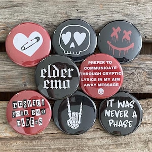 May include: A collection of round, pin-back buttons with various designs and text. The buttons feature a heart with a safety pin, a skull with heart eyes, a smiley face with dripping blood, and the words "elder emo". Other buttons say "Respect Your Emo Elders" and "It Was Never A Phase".