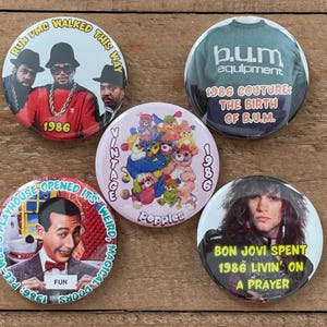May include: A collection of five vintage pinback buttons. The buttons feature images of Run DMC, Bon Jovi, Pee-wee Herman, and B.U.M. Equipment. The buttons have text that reads "1986" and other phrases.