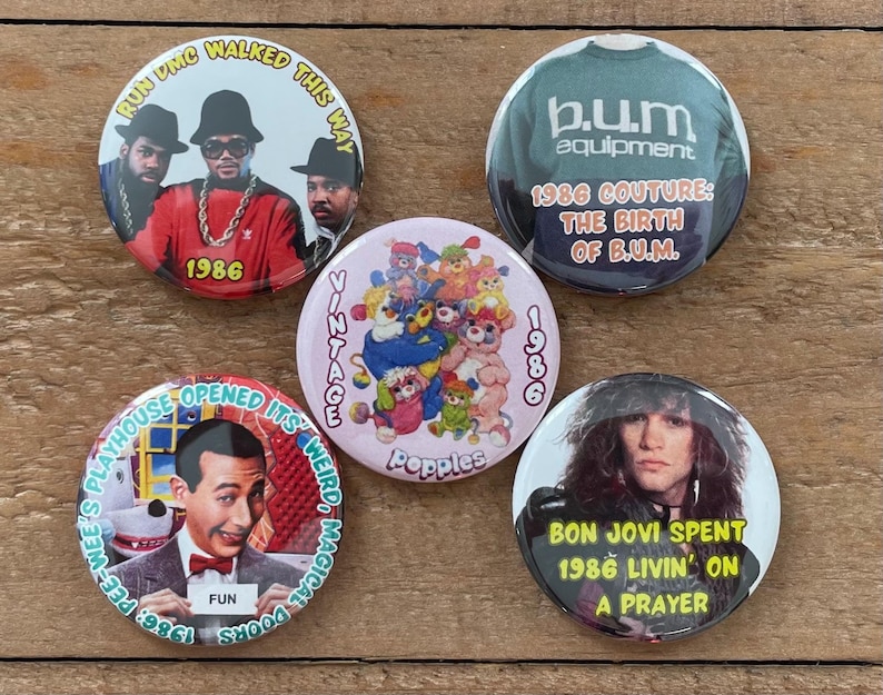 May include: A collection of five vintage pinback buttons. The buttons feature images of Run DMC, Bon Jovi, Pee-wee Herman, and B.U.M. Equipment. The buttons have text that reads "1986" and other phrases.