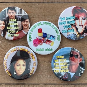 May include: A collection of five round buttons with images and text from the 1980s. One button features Ferris Bueller, another Cyndi Lauper, and a third Janet Jackson. The other two buttons have text that reads "Don't push me, push a push pop" and "We felt the need, the need for speed."