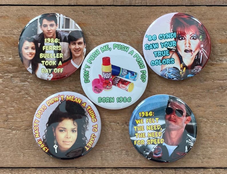 May include: A collection of five round buttons with images and text from the 1980s. One button features Ferris Bueller, another Cyndi Lauper, and a third Janet Jackson. The other two buttons have text that reads "Don't push me, push a push pop" and "We felt the need, the need for speed."