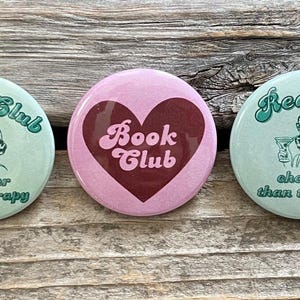 Pode incluir: Tr&ecirc;s bot&otilde;es redondos com um design vintage. Dois bot&otilde;es s&atilde;o verde claro com as palavras "Book Club" e "Reading" em uma fonte cursiva, uma ilustra&ccedil;&atilde;o retr&ocirc; e a frase "cheaper than therapy". O bot&atilde;o central &eacute; rosa com um cora&ccedil;&atilde;o bord&ocirc; e as palavras "Book Club".